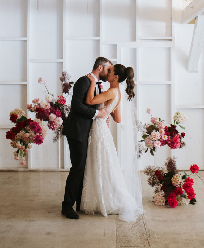 Happy couple sharing a kiss in the ceremony space at Gather and Tailor, a luxury Melbourne wedding venue, surrounded by stunning red and pink flowers by Anatomy of Flowers. The elegant floral design creates a romantic and timeless atmosphere.