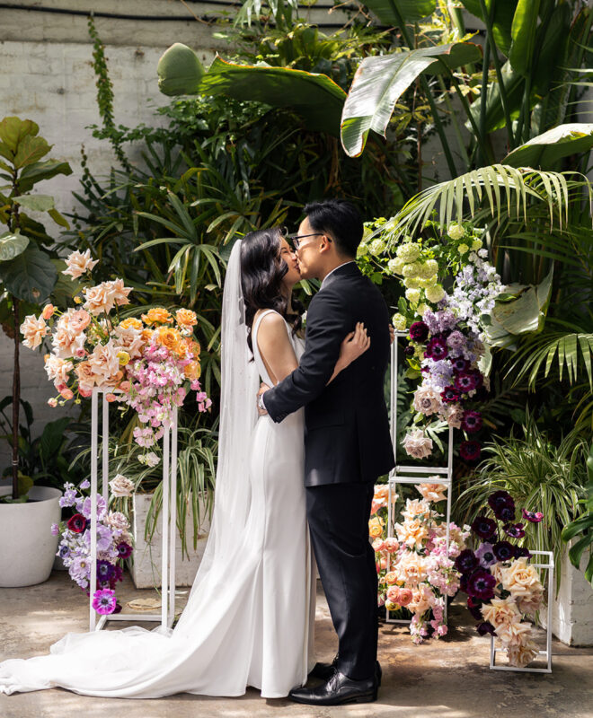 The couple shares a tender kiss in the picturesque Garden Room at The Button Factory, a one-of-a-kind Melbourne venue that blends lush greenery with a sophisticated, romantic atmosphere.