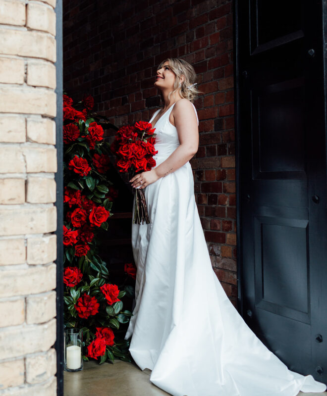 Beautiful bride holding a bouquet of vibrant red roses, with Chapel 1885's signature staircase elegantly adorned with lush red roses by Anatomy of Flowers, creating a romantic and sophisticated atmosphere for a Melbourne wedding celebration.