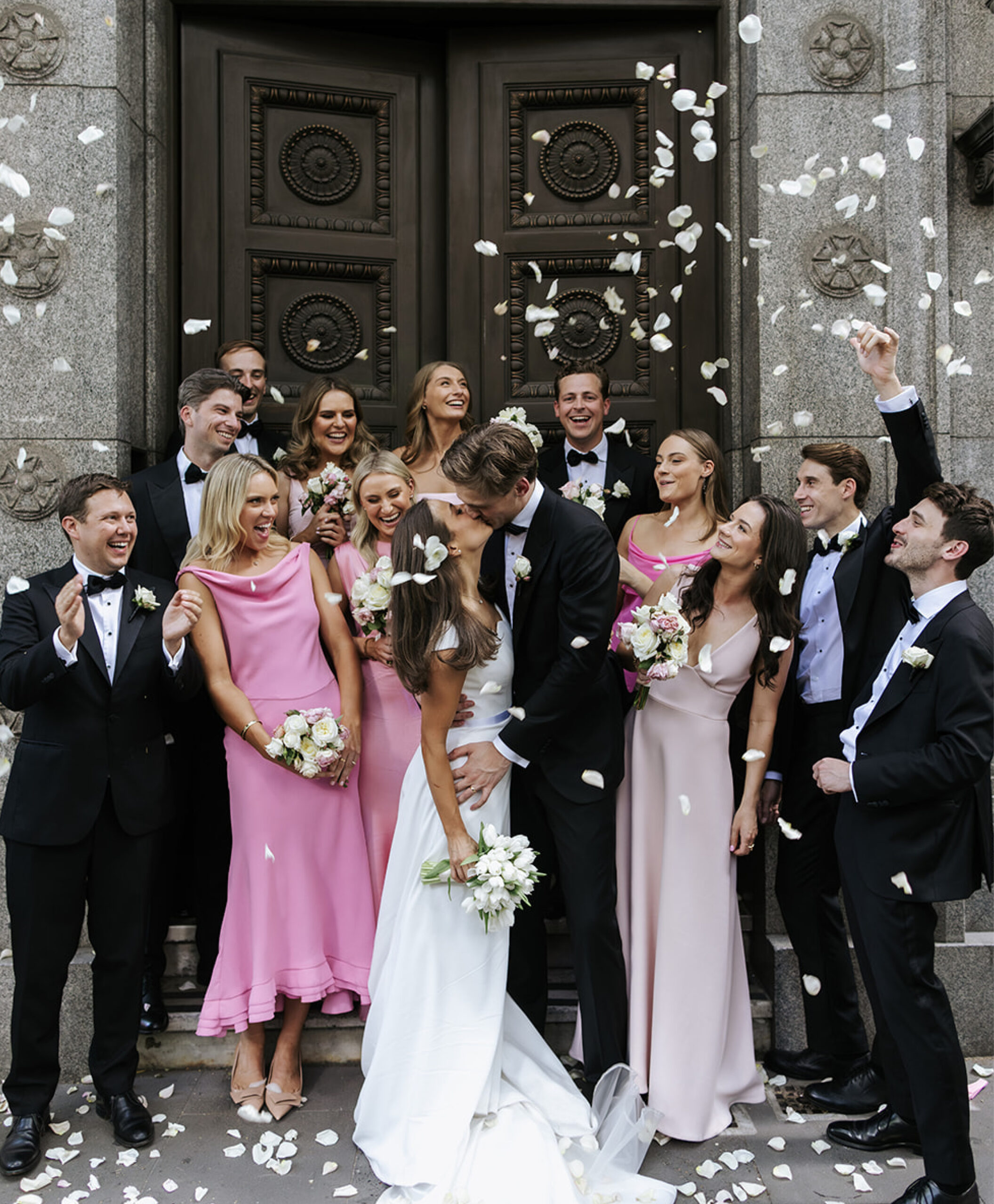 Surrounded by loved ones, a couple shares a magical kiss in the elegant doorway of The Trust’s iconic revolving door, capturing a timeless moment at this premier Melbourne venue for weddings and events.