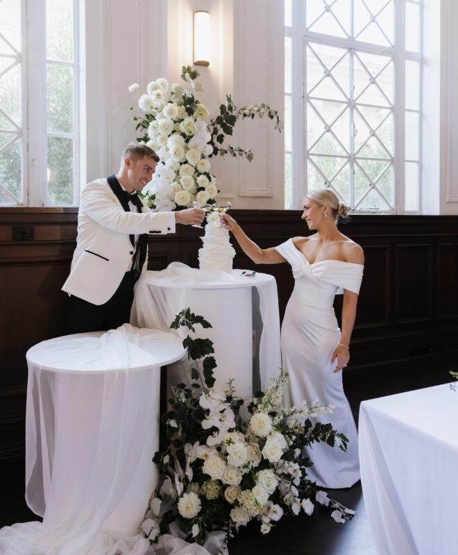 The happy couple cutting their wedding cake in The Trust Melbourne’s Grand Hall, surrounded by stunning white roses by Anatomy of Flowers, creating an elegant and unforgettable moment for their wedding celebration.